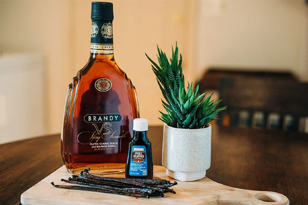 A bottle of brandy, almond extract, a plant, and vanilla beans on a table